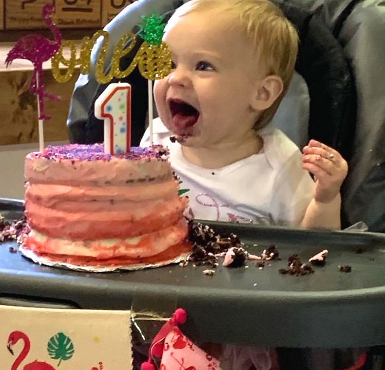 Baby sits in high chair and smiles behind cake with candle.