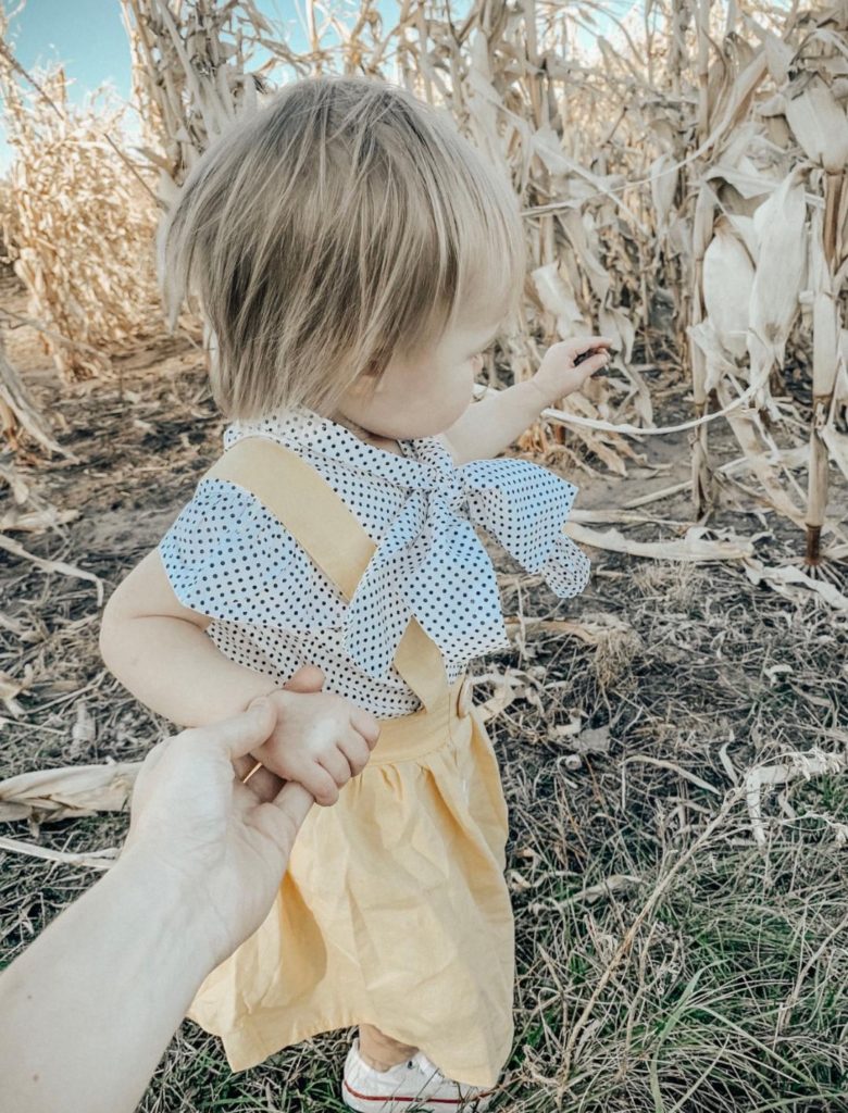 Girl in golden yellow jumper dress pulls mother towards corn maze.